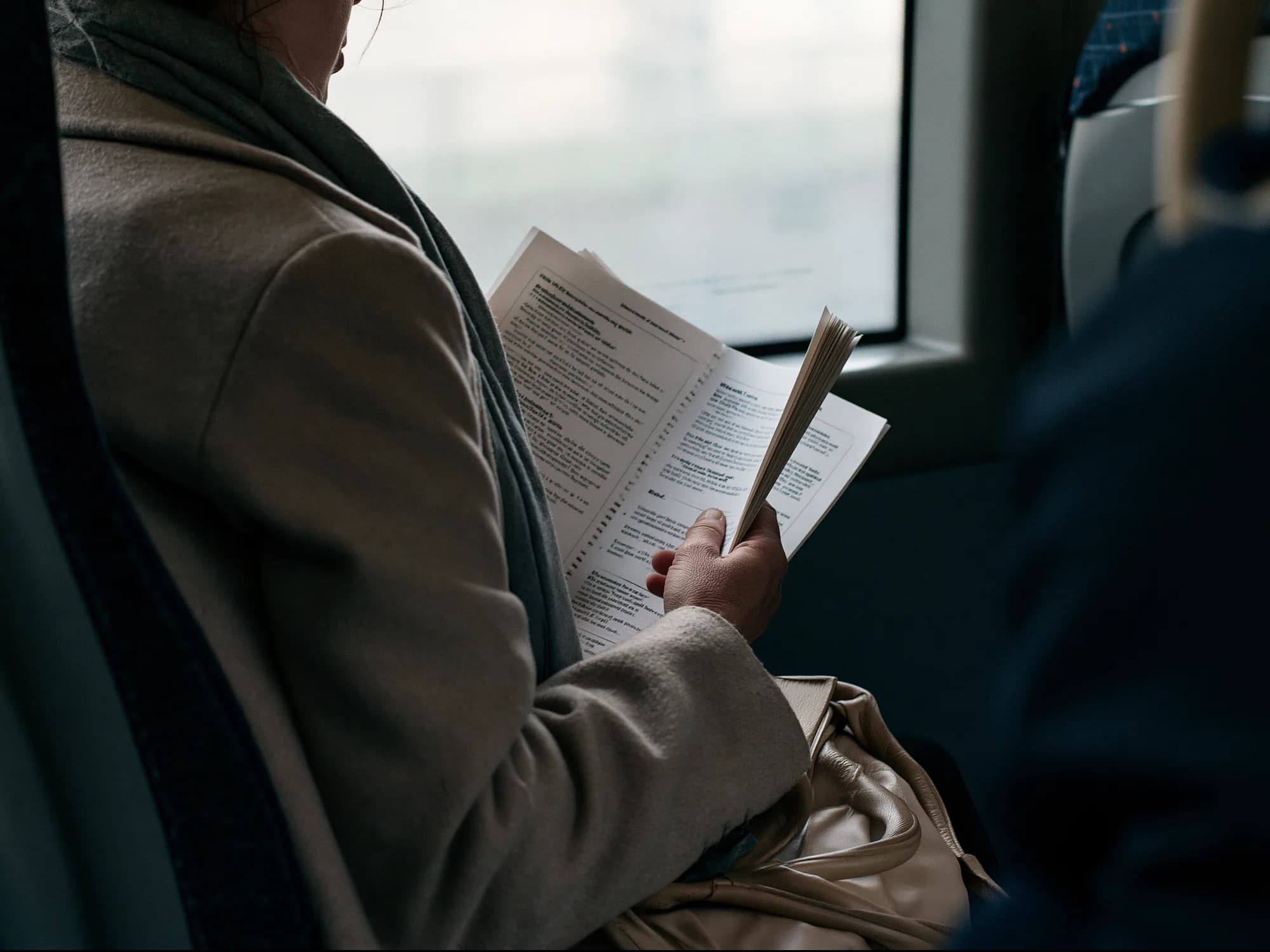 A person on a bus reading a stapled printed guide, seen over the shoulder, grey daylight through the window