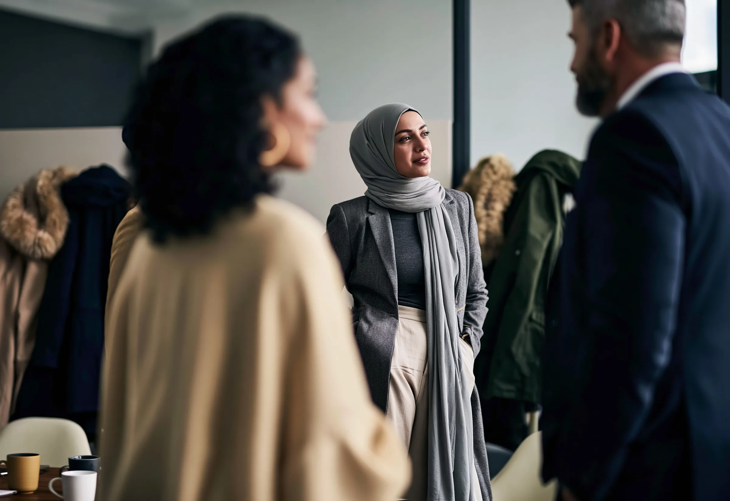 Three colleagues standing in conversation after a meeting, coats still on the rack behind them