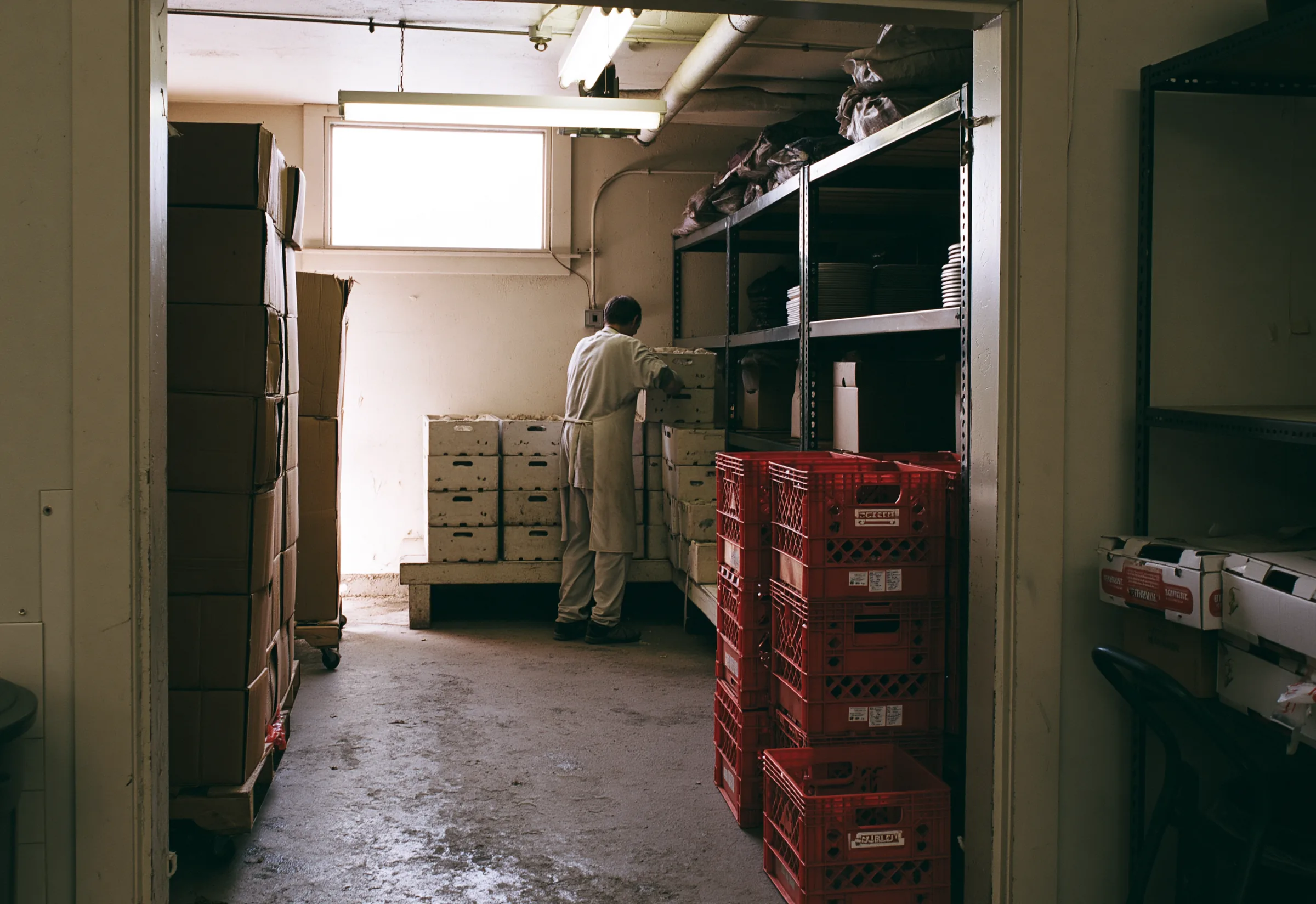 A worker in a pale coat and apron, seen from behind, moving a crate in a small food storage room stacked with boxes and crates