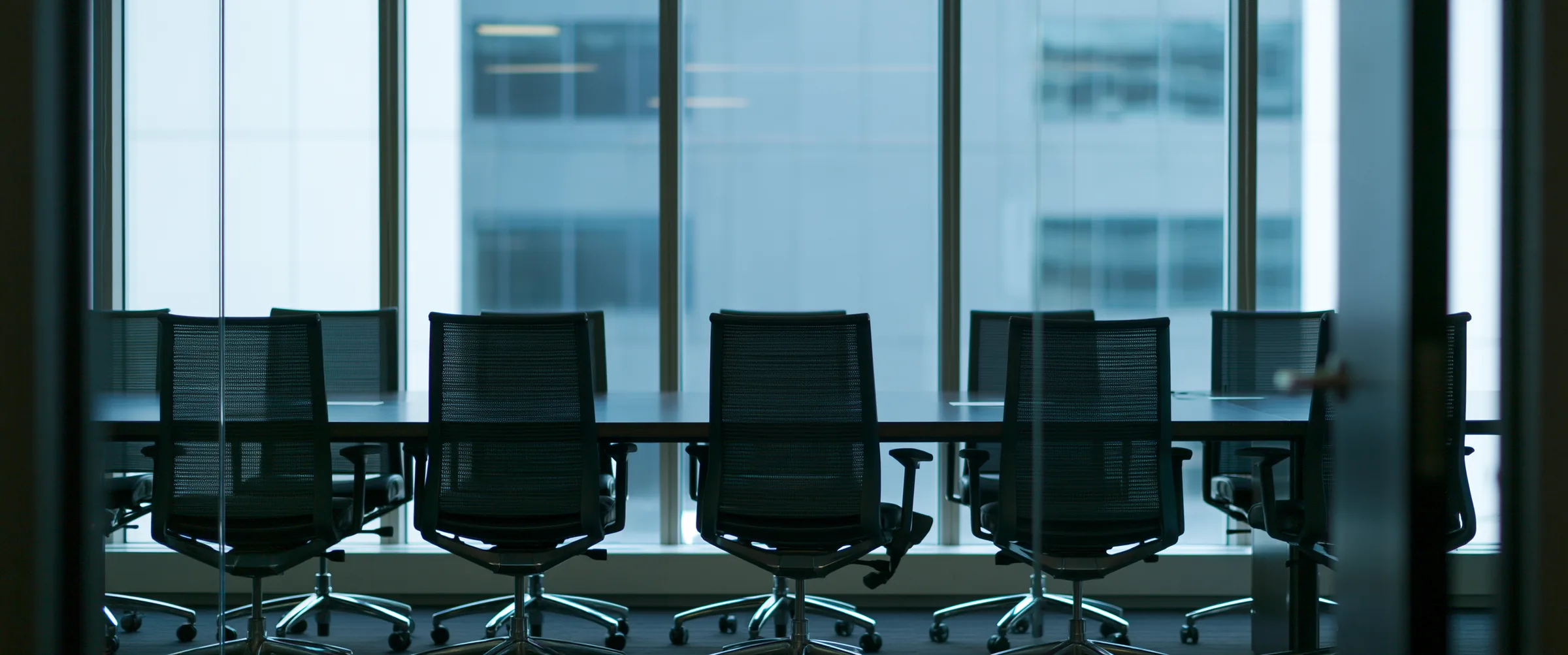 An empty boardroom seen through glass, chairs ranged along the table at a wall of windows