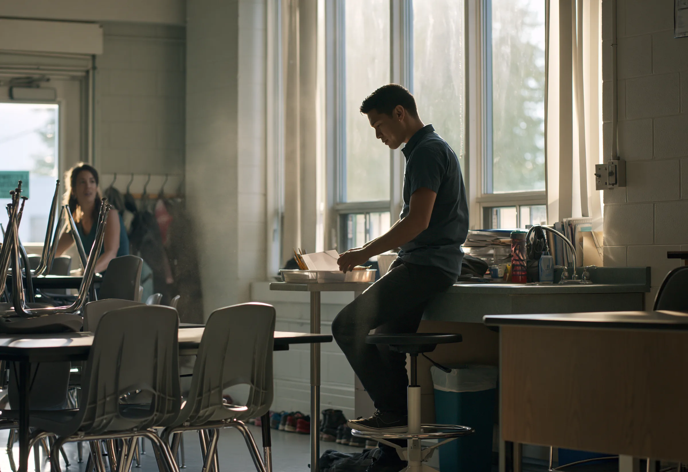 A teacher working through papers at a high table in an empty classroom in late afternoon light, a colleague at the far end of the room