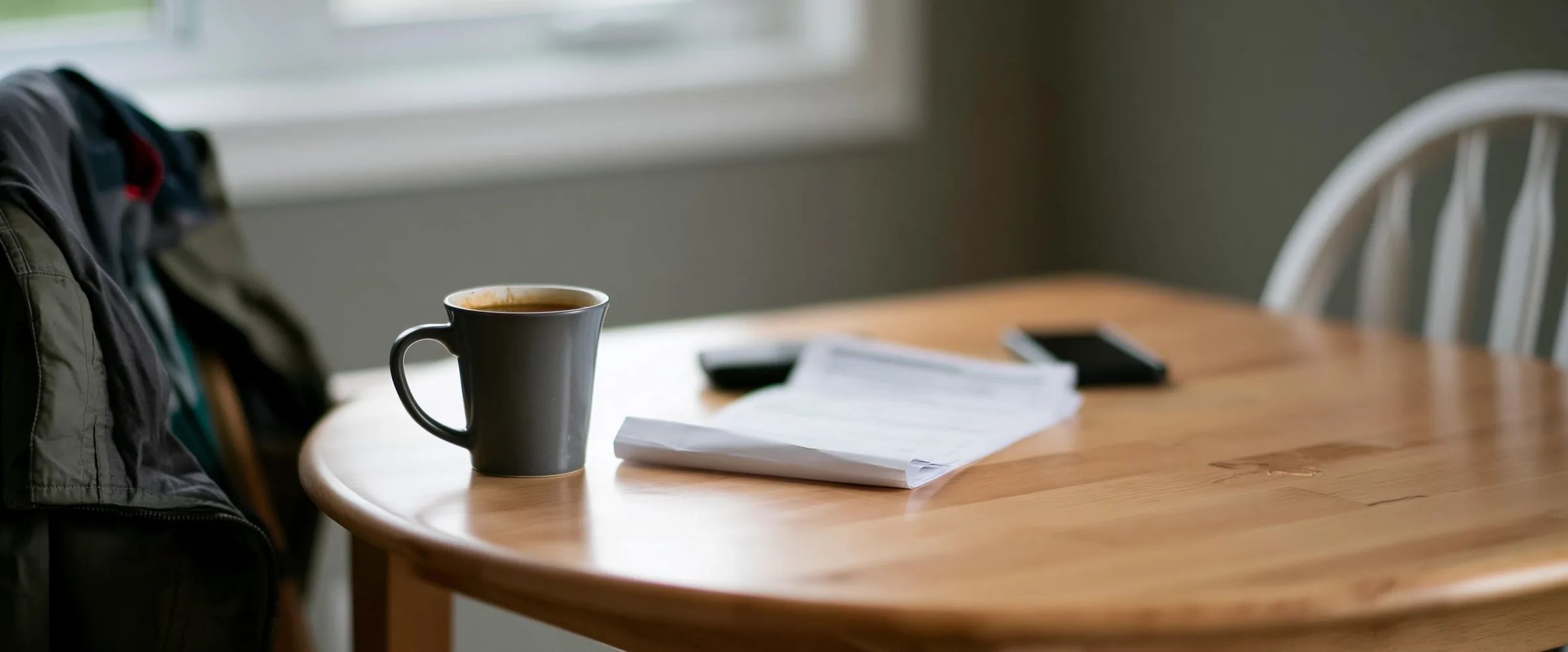A mug of tea, a folded paper form and a face-down phone on a kitchen table in weekday morning light, a jacket over the chair back