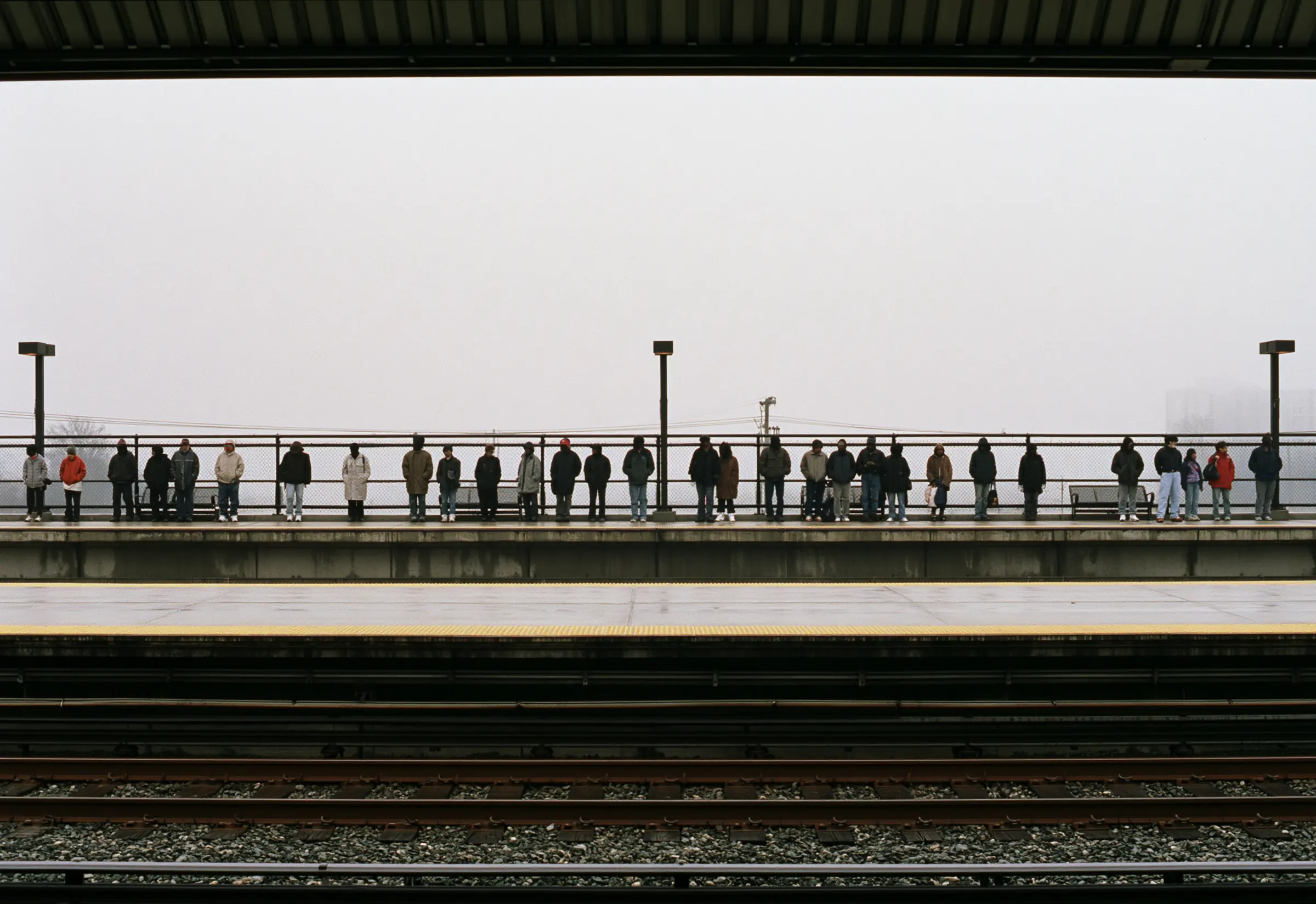 About thirty people waiting in a row along a railway platform under a pale overcast sky, none of them identifiable