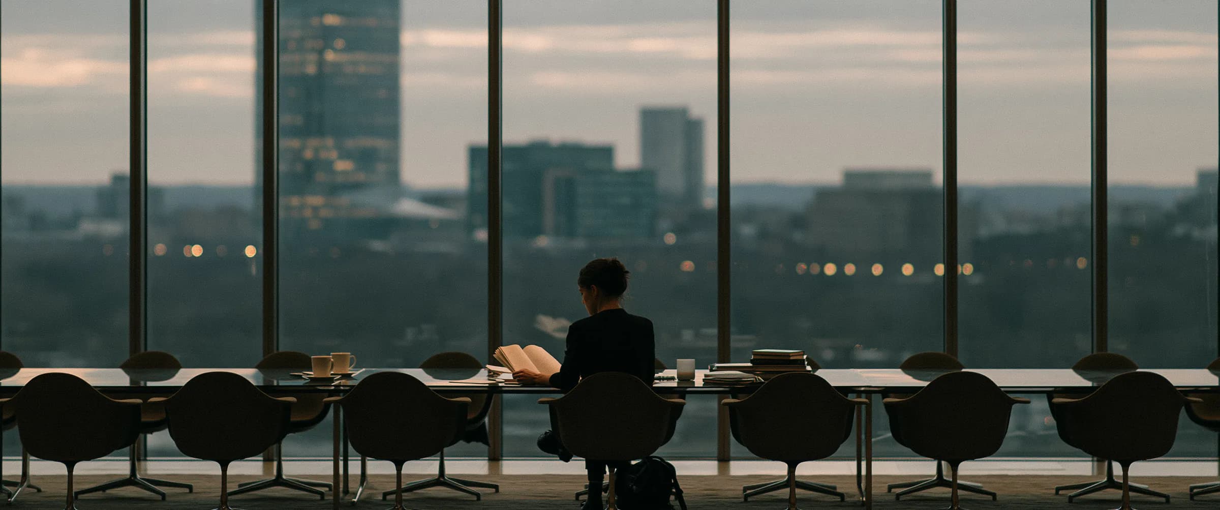 One person alone at a long table at dusk, reading, the city beyond the glass behind them